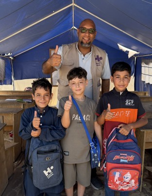 Thre students with back to school bags, with their thumbs up. Behind, a HA staff member, smiling and with his thumb up. 